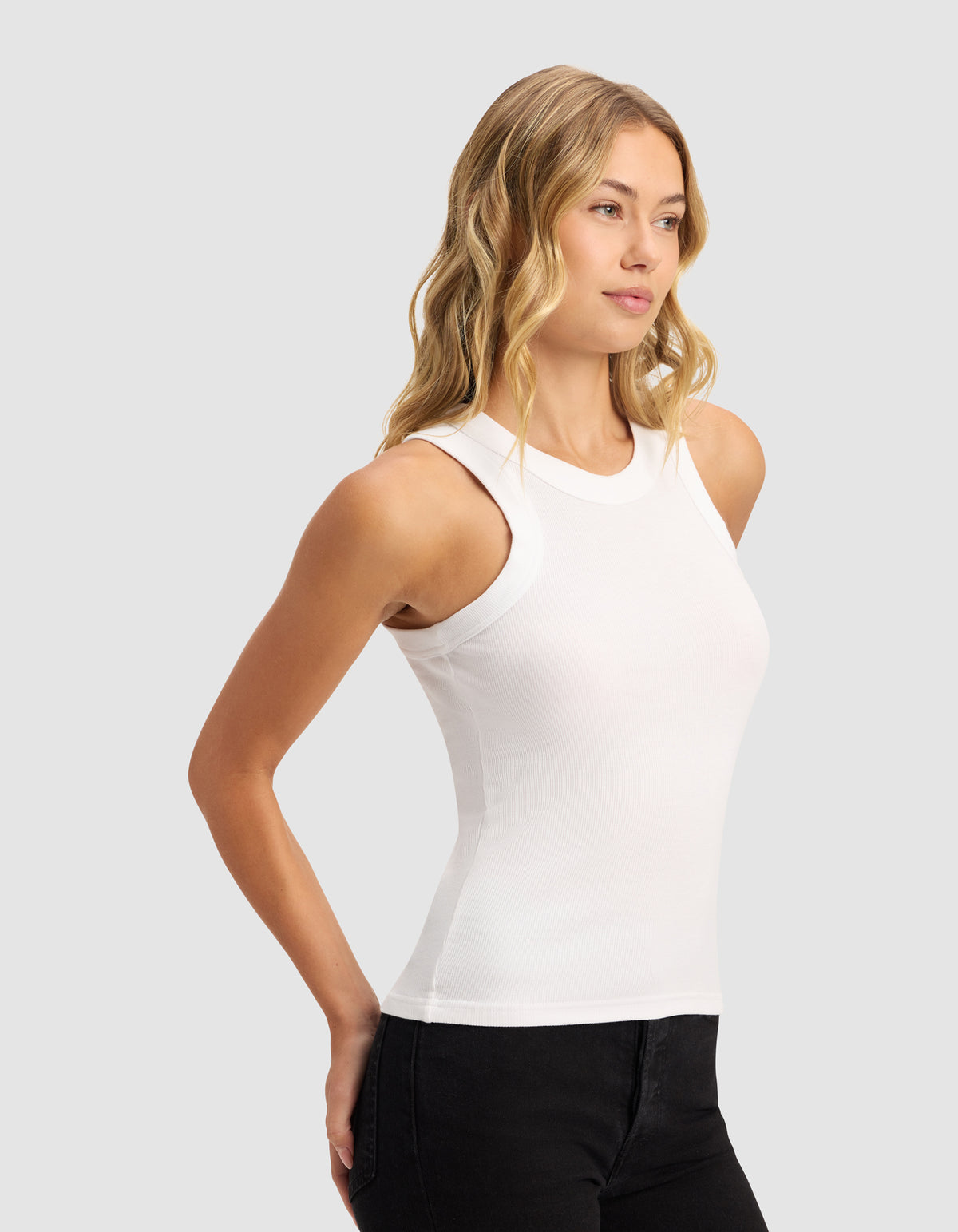 A woman with wavy blonde hair wears the Cozy Earth Women's Fine Ribbed Tank in white and black pants, standing against a plain light gray background, looking slightly to the side with her hands behind her back.