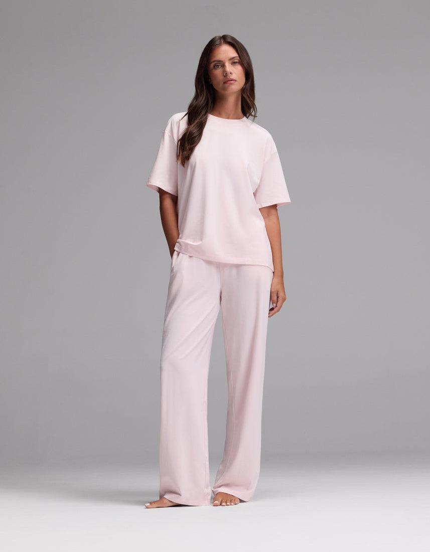 A woman stands barefoot against a plain gray background, wearing the Cozy Earth Women’s First Layer Pajama Short Sleeve Tee & Pajama Pant Set in Ballet—a loose-fitting, light pink ensemble featuring short sleeves and wide-leg pants.