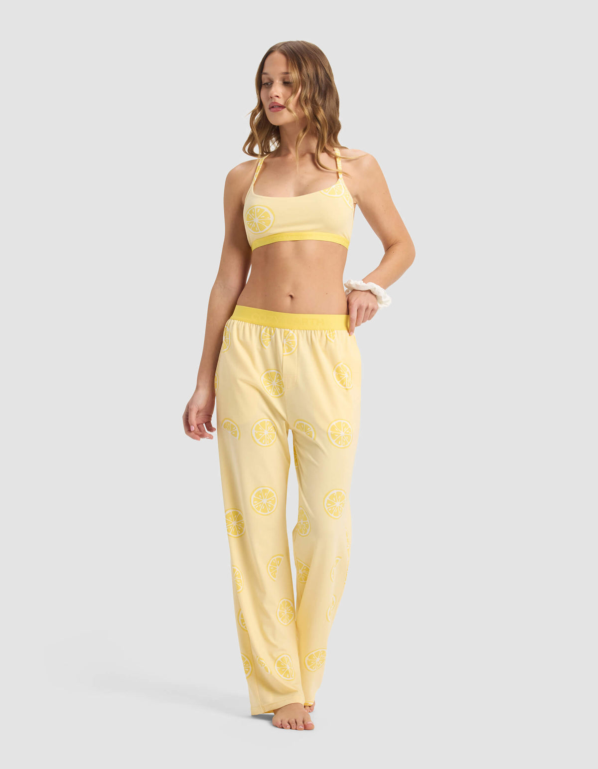 A woman stands barefoot against a plain background wearing Cozy Earth's Women's First Layer Pajama Pant, featuring a yellow color and lemon slice pattern. The set includes a crop top and loose-fitting pajama pants.