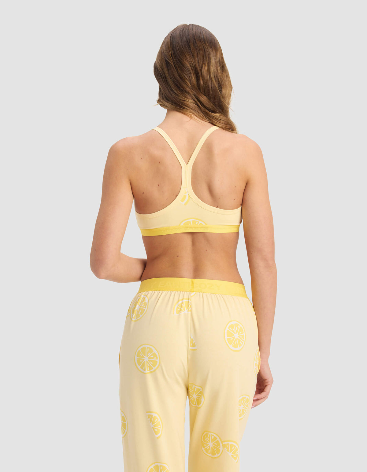 A woman with long, wavy hair models the Cozy Earth Women's First Layer Pajama Soft Bra and matching lemon-print pajama pants, standing with her back to the camera against a light gray background.