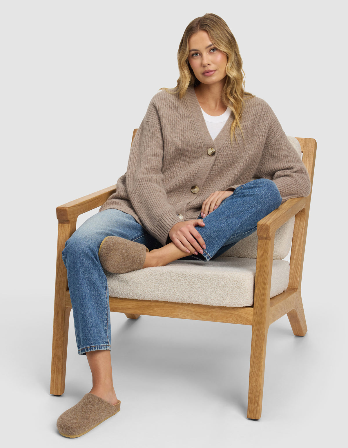 A woman with long blonde hair sits relaxed on a wooden armchair, wearing blue jeans and Cozy Earth’s Lakehouse Clog slippers. One leg is crossed over the other, and she looks calmly at the camera.