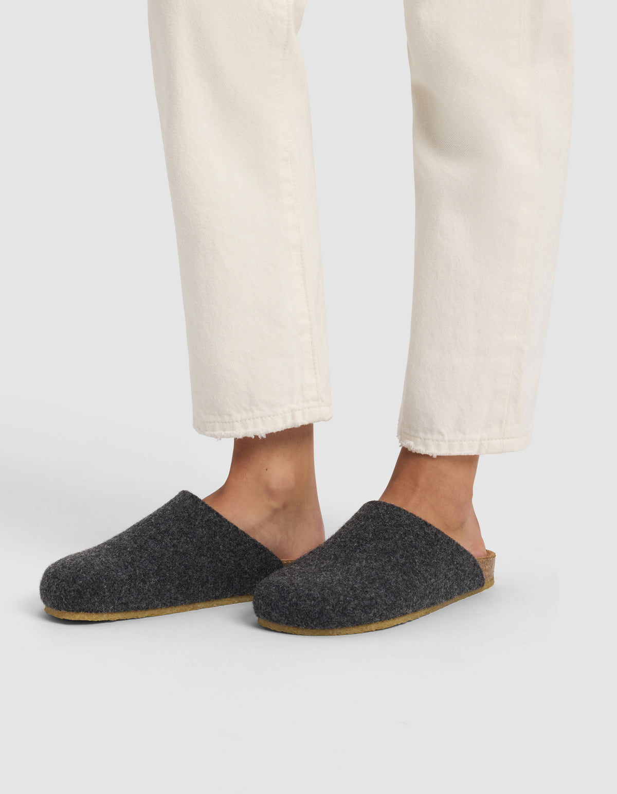 A person wearing off-white pants and Cozy Earth’s Lakehouse Clog—dark gray wool slippers with light brown soles—is standing against a plain light background.|Color:Graphite