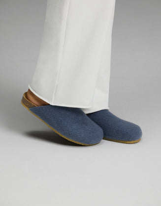 A person wearing white pants and Cozy Earth Lakehouse Clog slippers in light blue, featuring cork soles, stands on a light gray surface.|Color:Light Blue