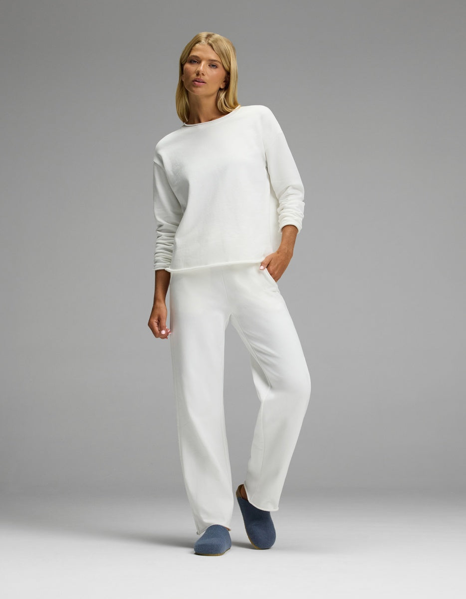 A woman stands in a relaxed pose wearing an all-white long-sleeve top and matching pants, with one hand in her pocket, against a plain gray background. She completes the look with Cozy Earth’s Lakehouse Clog slippers.|Color:Light Blue