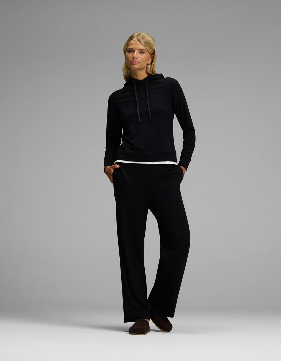 A woman stands with hands in her pockets, wearing the Lakehouse Clog in Suede by Cozy Earth, paired with a black long-sleeve hoodie and matching black pants layered over a white shirt, against a plain gray background.