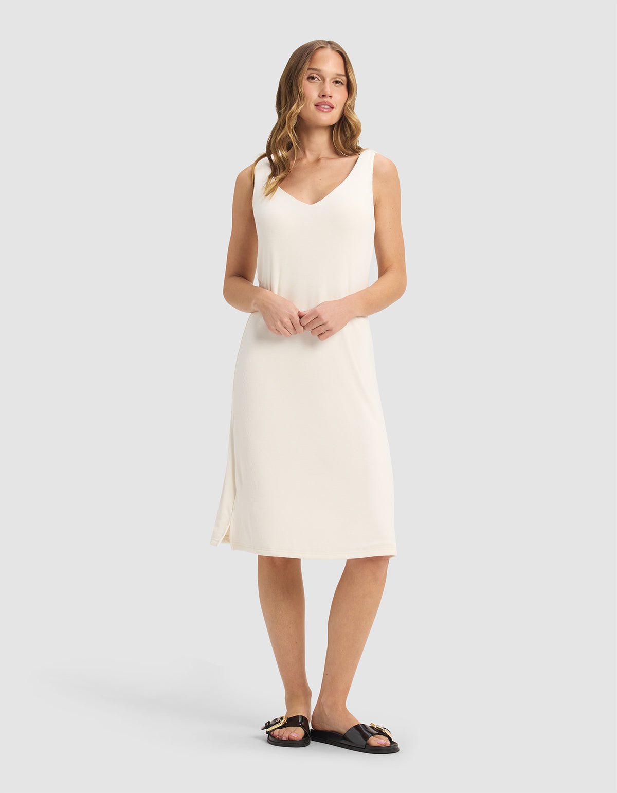 A woman stands against a light gray background, wearing the Cozy Earth Women’s Bamboo Rib-Knit V-Neck Tank Lounge Dress in Ivory. She has loose, wavy hair and pairs the knee-length, sleeveless dress with black slip-on sandals featuring gold accents.