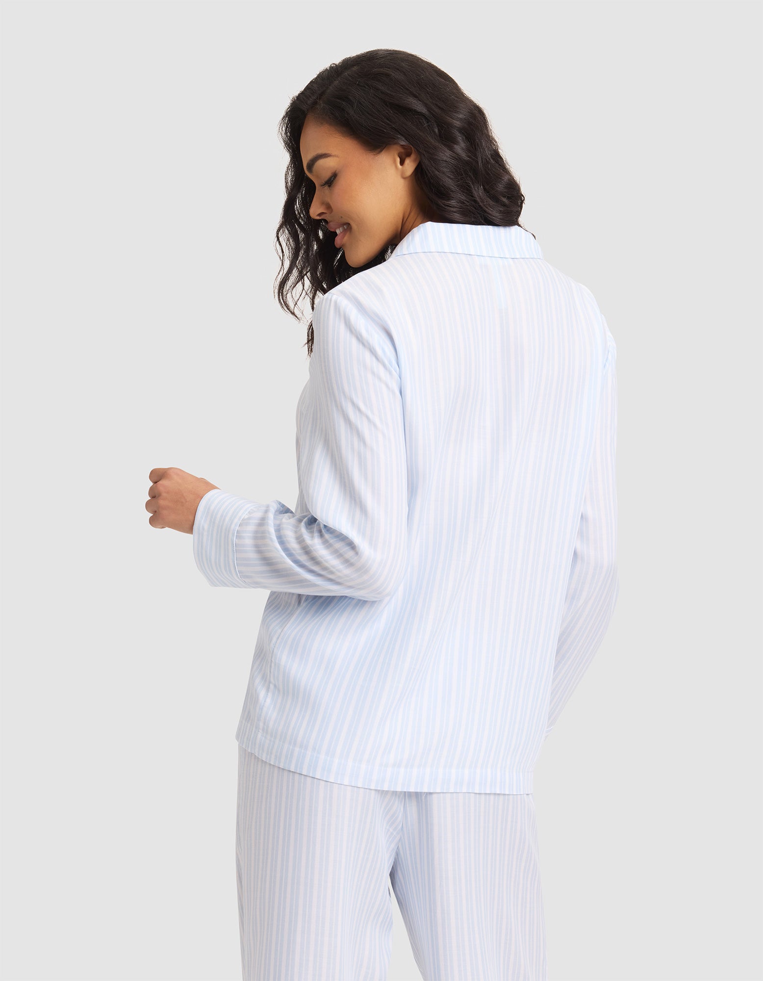 A woman with wavy hair smiles and looks down, shown from the back and slightly turned to the side, wearing the Cozy Earth Women's Soft Woven Long Sleeve Pajama Top in light blue and white stripes against a plain light gray background.|Color:Spring Blue Stripe