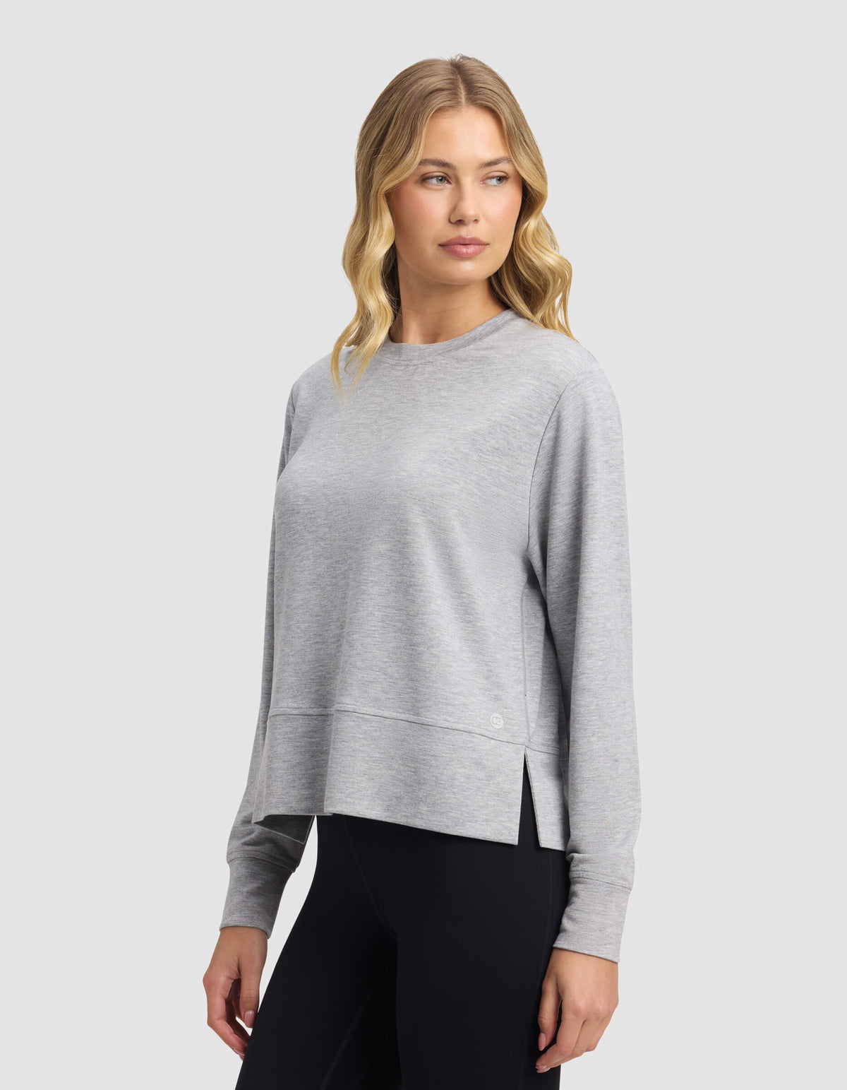 A woman with wavy blonde hair wears the Cozy Earth Women's StudioLite Crewneck in light gray with black leggings, standing against a plain light gray background and looking slightly to her left.