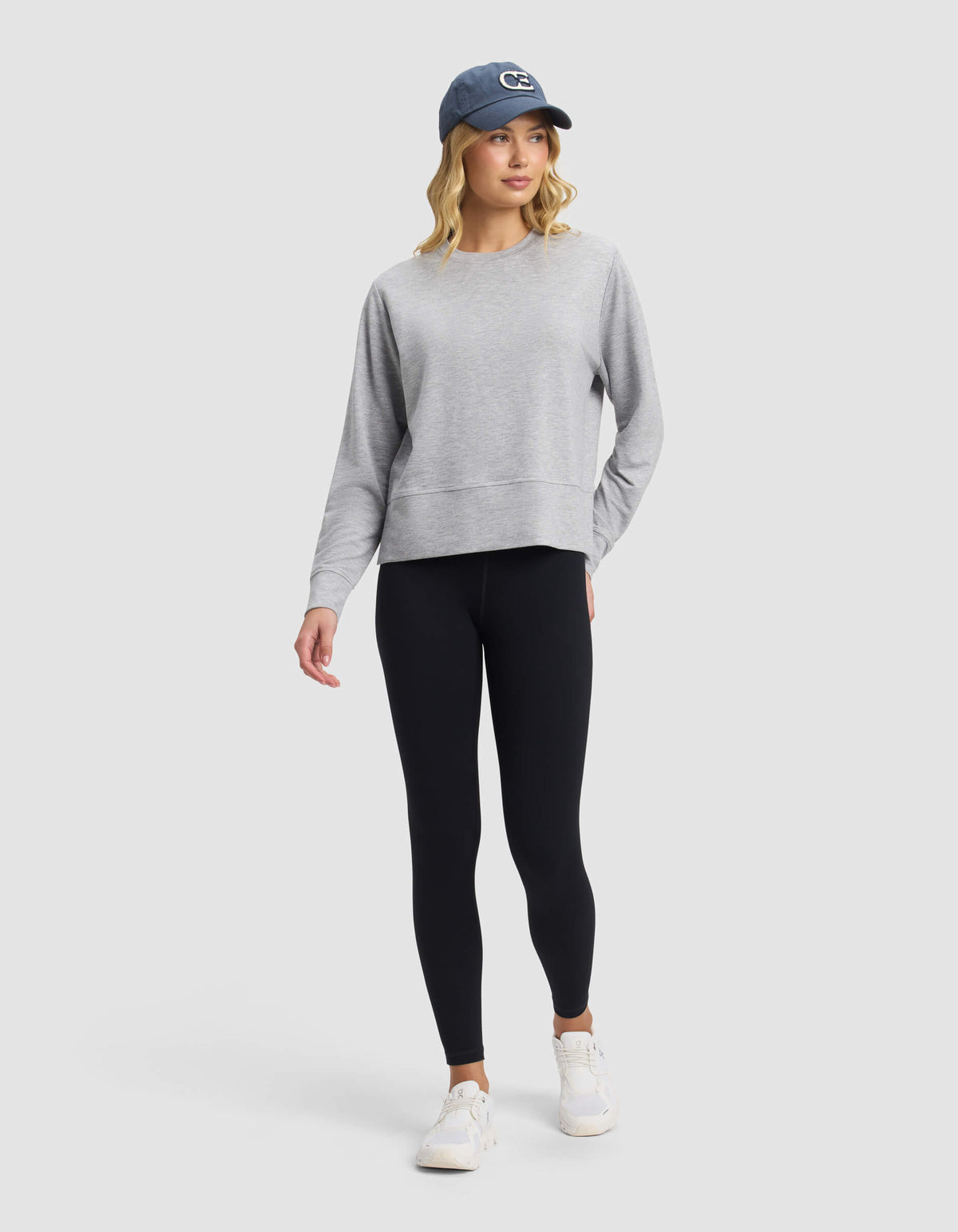 A woman wears the Cozy Earth Women's StudioLite Crewneck, paired with black leggings, white sneakers, and a blue baseball cap. She stands against a light gray background, looking slightly to the side with one hand in her pocket.