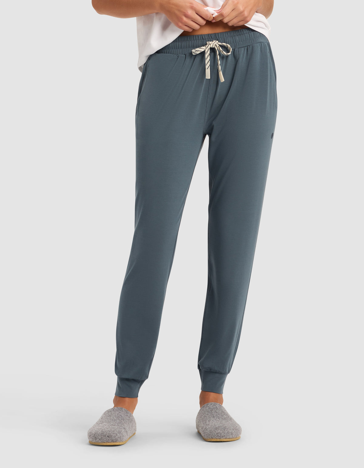 A person wearing Cozy Earth's Women's Studio Jogger in teal with a striped drawstring, paired with a white top and gray slippers, stands against a plain light background. Their face is not visible.