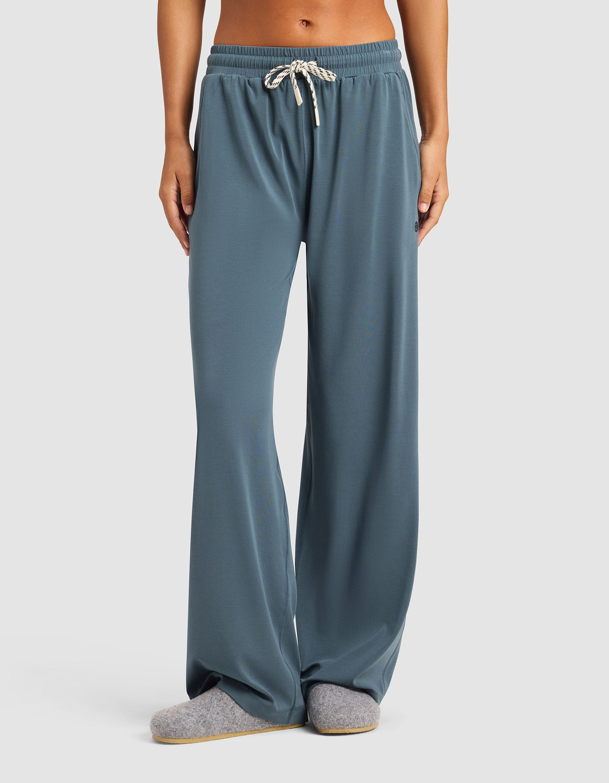 A person wears Cozy Earth's Women's Studio Wide Leg Pant in blue-gray, paired with gray slippers, standing against a plain light background. Only the lower half of the body is shown.