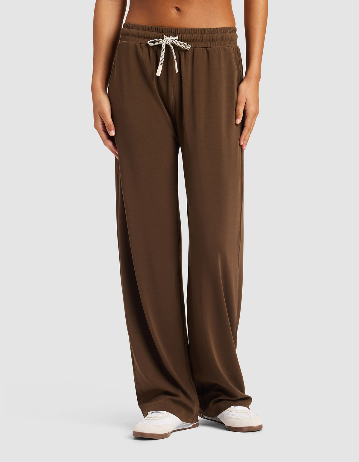 A person is standing against a light gray background wearing Cozy Earth's Women's Studio Wide Leg Pant in brown with white sneakers, hands relaxed by their sides. The pants feature a loose fit and drawstring waist.