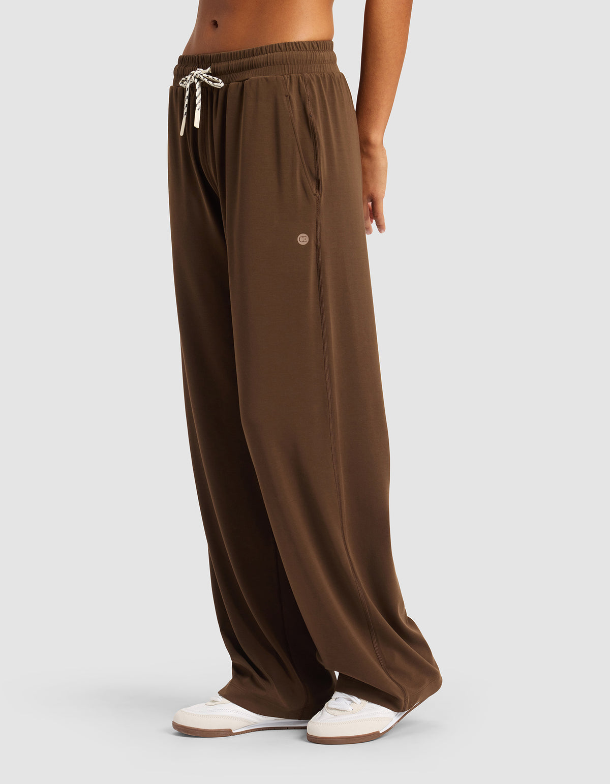 A woman wears Cozy Earth Women's Studio Wide Leg Pant—loose-fitting brown pants with an elastic waistband and white drawstring—paired with white sneakers, standing against a plain light gray background.