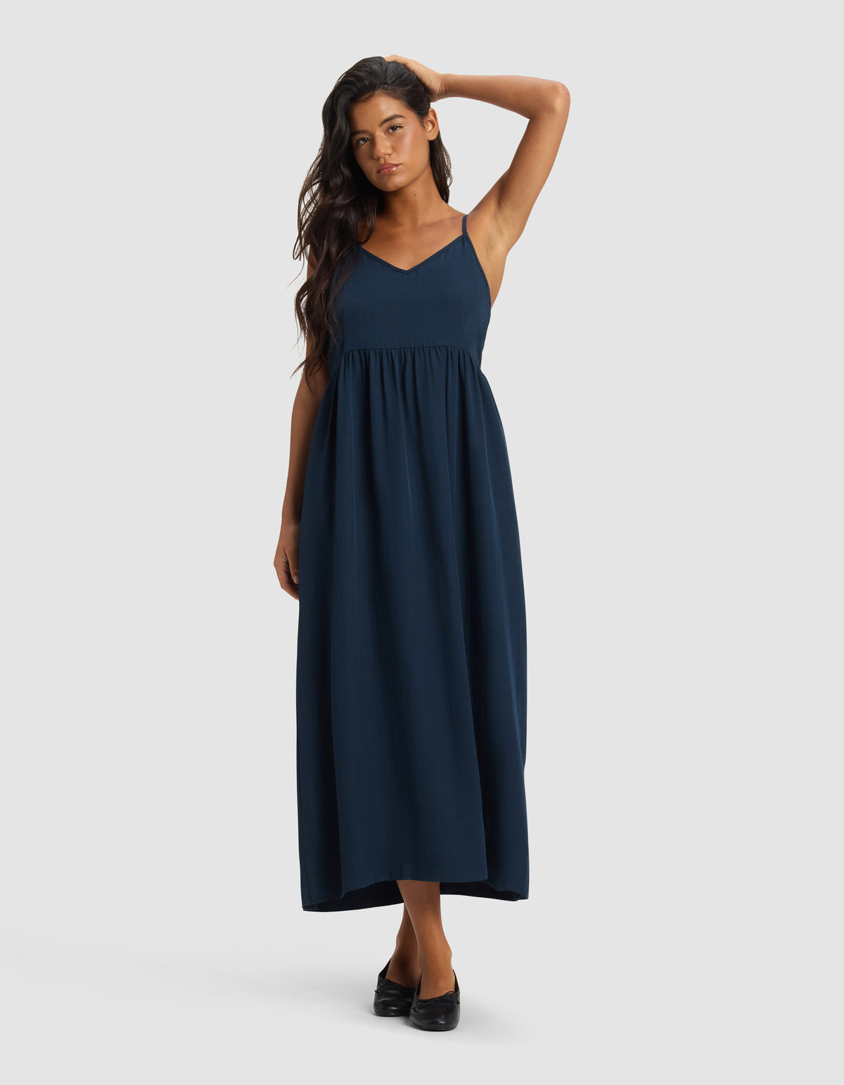 A woman poses on a plain background wearing Cozy Earth's Women's Sunset Gathered Dress - Last Chance, a sleeveless navy maxi with a V-neck and empire waist. She has long, dark hair and black shoes, touching her head with one hand.|Color:Eclipse