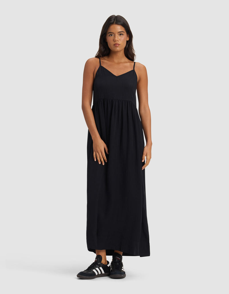 A woman faces forward in a Cozy Earth Women's Sunset Gathered Dress – Last Chance, an ankle-length, sleeveless black dress with thin straps, paired with black Adidas sneakers against a plain light gray background.