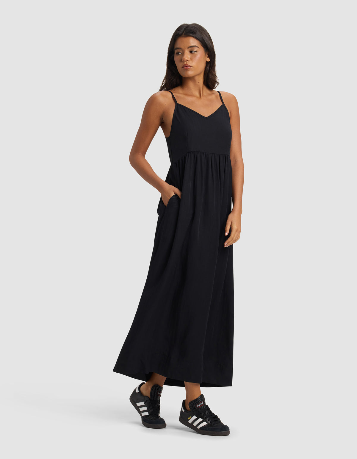 A woman wearing the Cozy Earth Women's Sunset Gathered Dress - Last Chance stands against a plain light gray background, looking to her left with one hand in her pocket and pairing the dress with black Adidas sneakers.