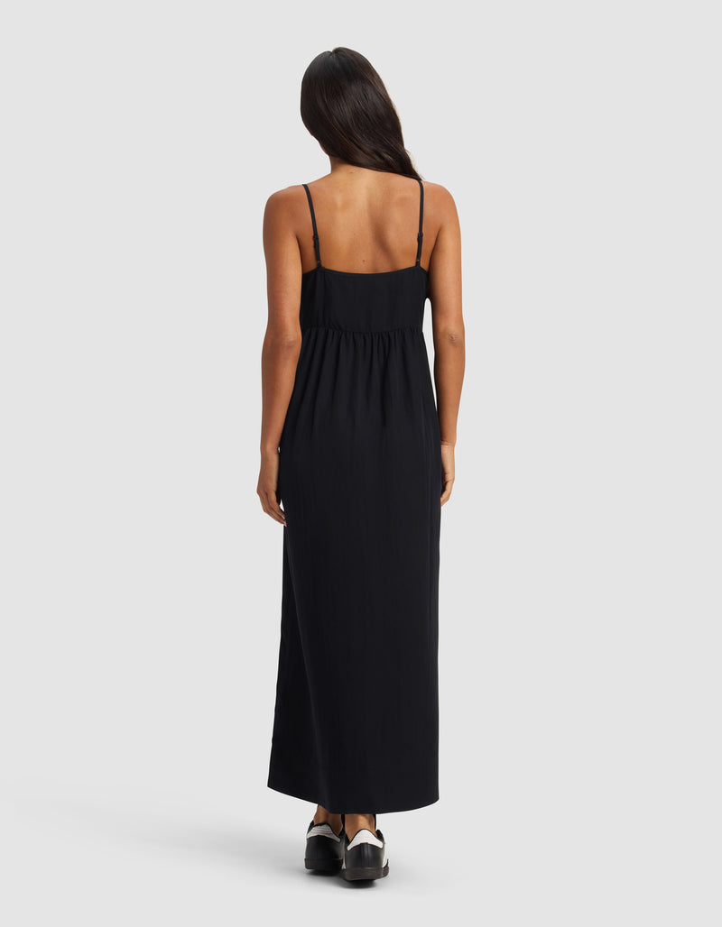 A woman with long dark hair stands facing away from the camera, wearing the Cozy Earth Women's Sunset Gathered Dress - Last Chance in black, styled with black shoes, set against a plain light background.