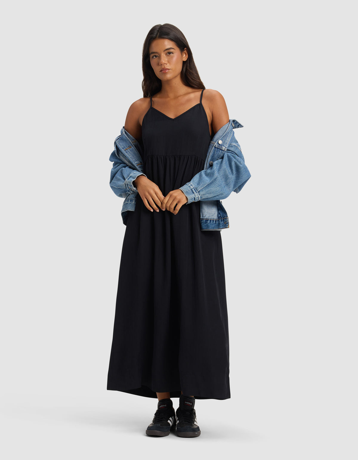 A woman with long dark hair and a neutral expression stands facing forward against a plain light grey background, wearing the Cozy Earth Women's Sunset Gathered Dress - Last Chance, with a blue denim jacket off her shoulders and black sneakers.