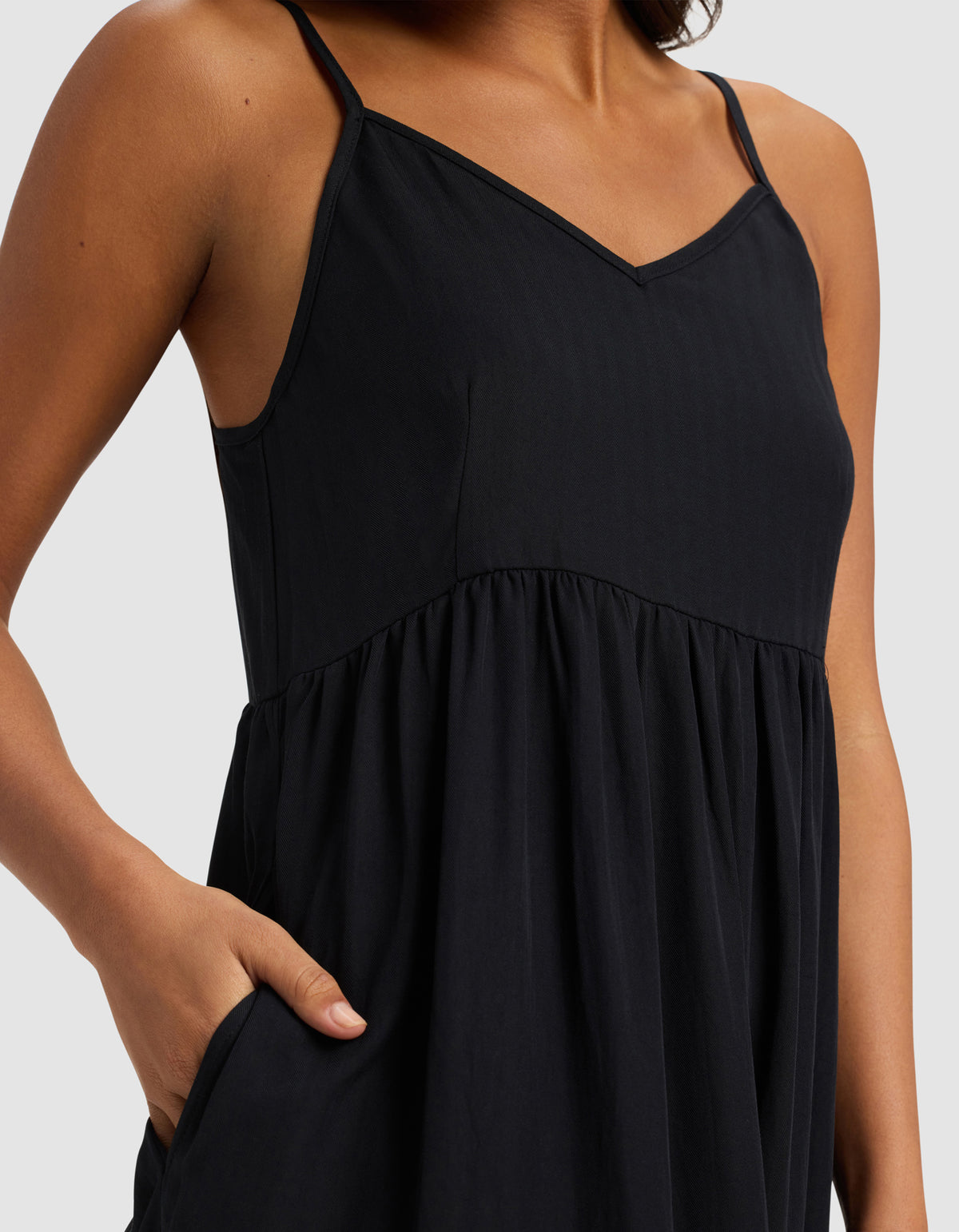 A model wears the Cozy Earth Women's Sunset Gathered Dress - Last Chance, a sleeveless black dress with thin straps and a V-neckline, shown from shoulders to mid-thigh, with one hand in the pocket.