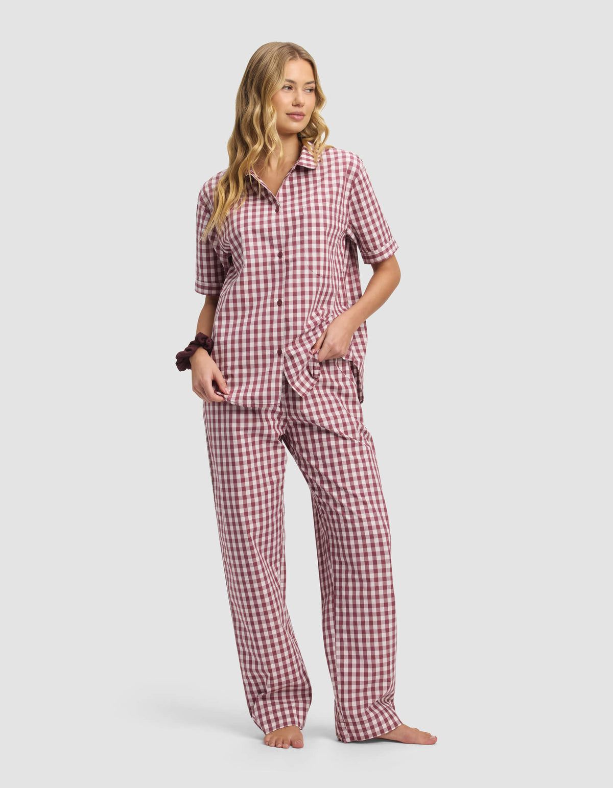 A woman with long, wavy blonde hair stands barefoot and relaxed, wearing Cozy Earth's Women's Sutton Pajama Pant in a maroon and white gingham print, paired with a matching short-sleeve, button-up top. One hand is in her pocket.