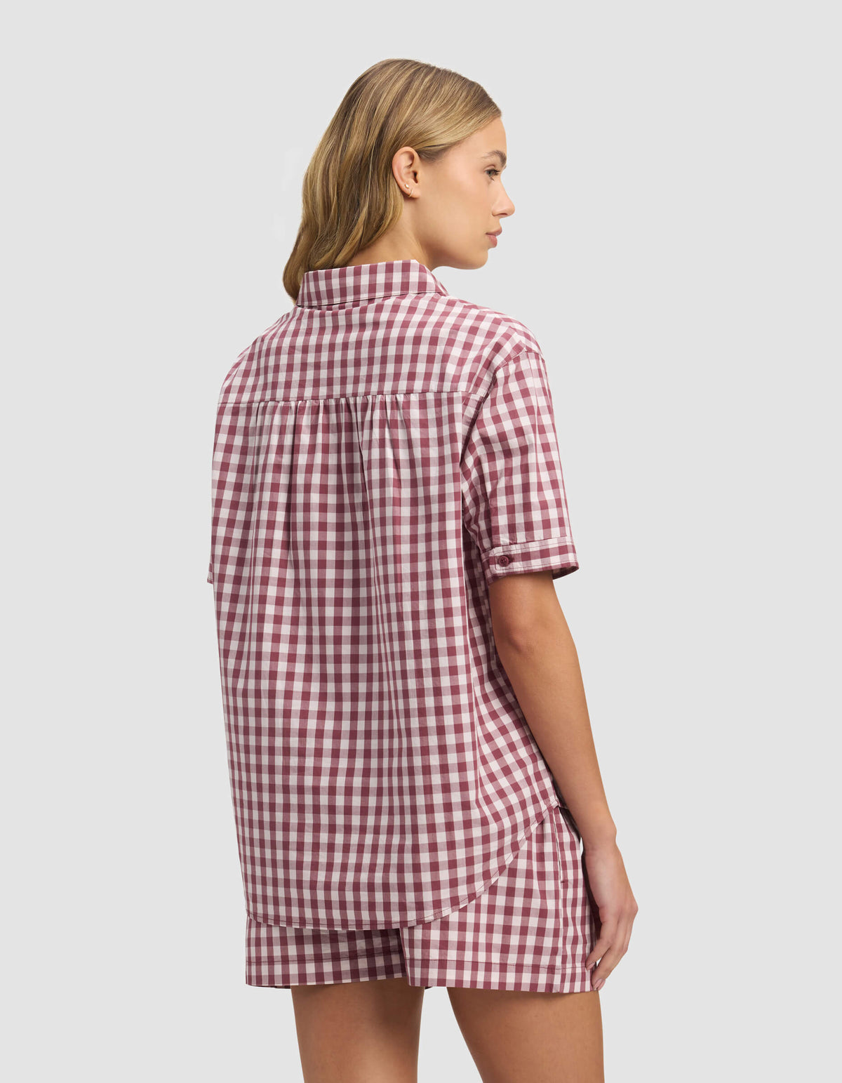 A woman with light brown hair, seen from behind against a plain light background, wears the Cozy Earth Women's Sutton Short Sleeve Pajama Top in an oversized red and white gingham pattern with matching shorts.