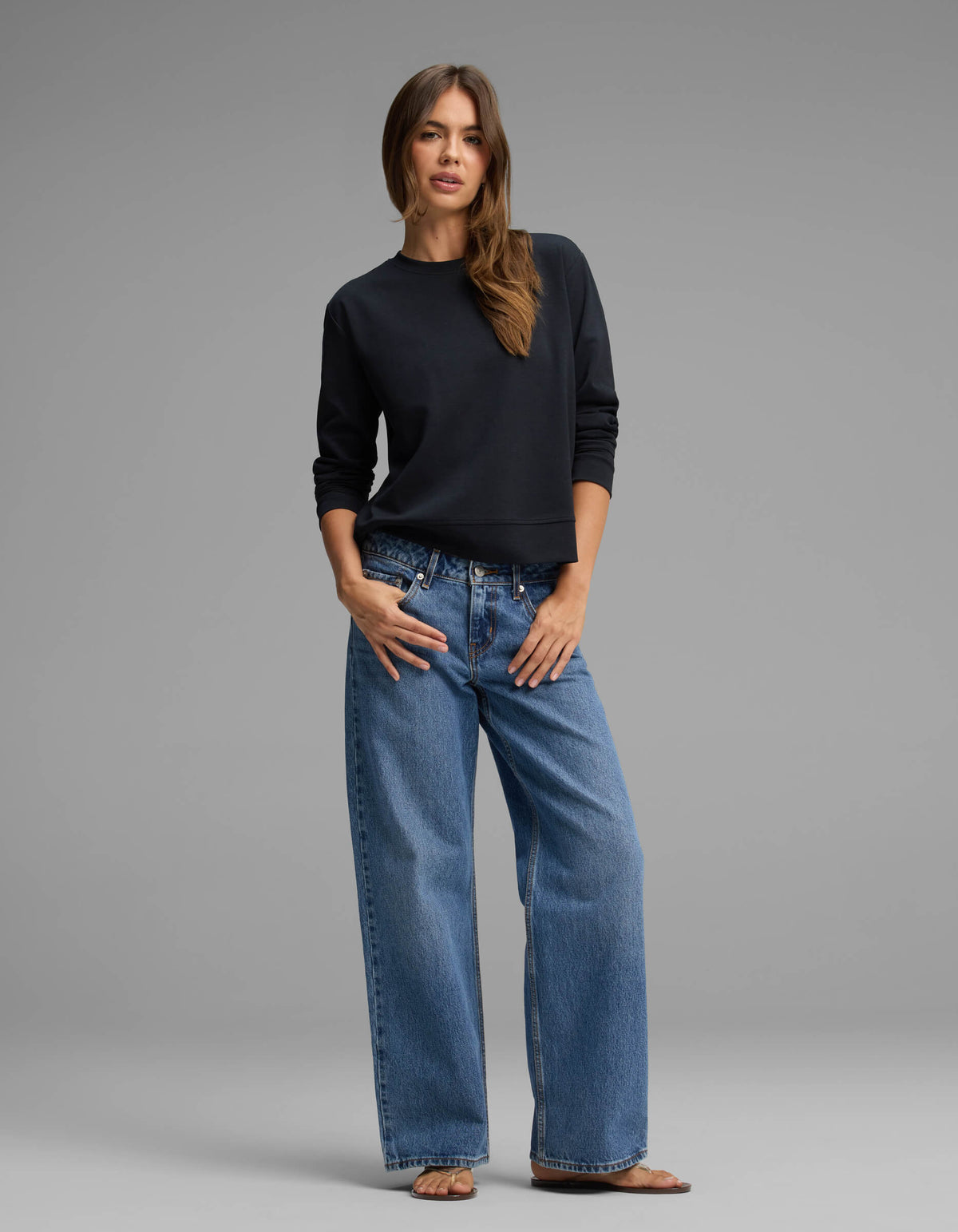 A woman with long brown hair stands against a gray background wearing the Cozy Earth Women's Versa Crewneck, paired with loose blue jeans and sandals. She poses with one hand in her pocket and the other resting by her side.