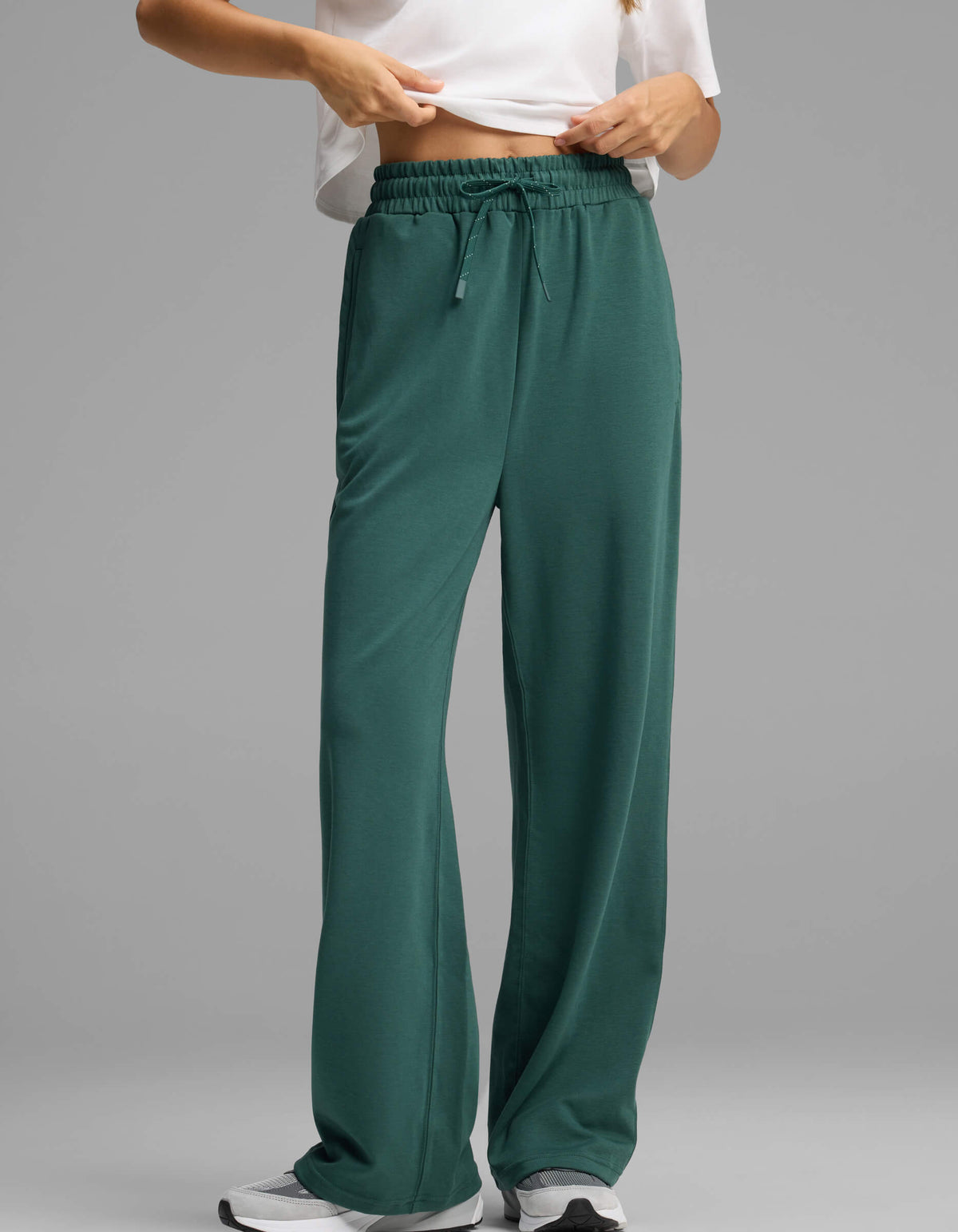 A model showcases the lower half of the Women's Versa Straight Leg Pant by Cozy Earth in green, paired with a white shirt and white sneakers, standing against a plain grey background and seen adjusting the waistband.