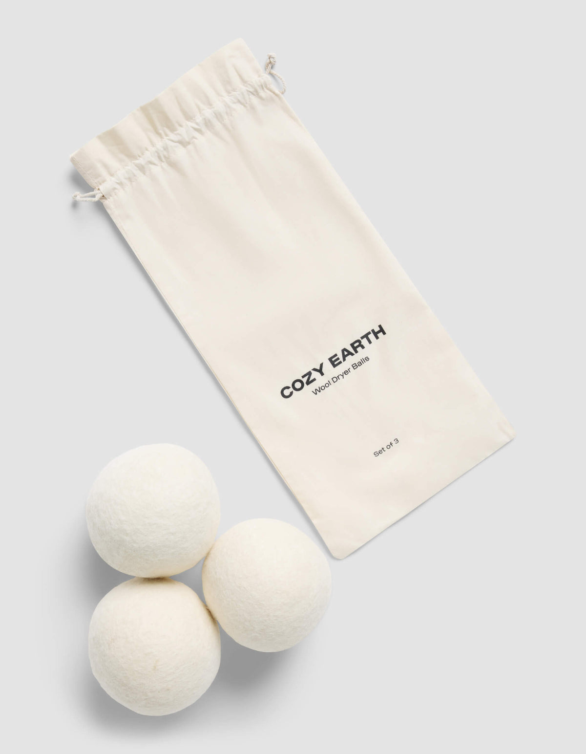 A cream-colored drawstring bag with "Cozy Earth" branding and "Wool Dryer Balls" label sits beside three white wool dryer balls on a light gray background.
