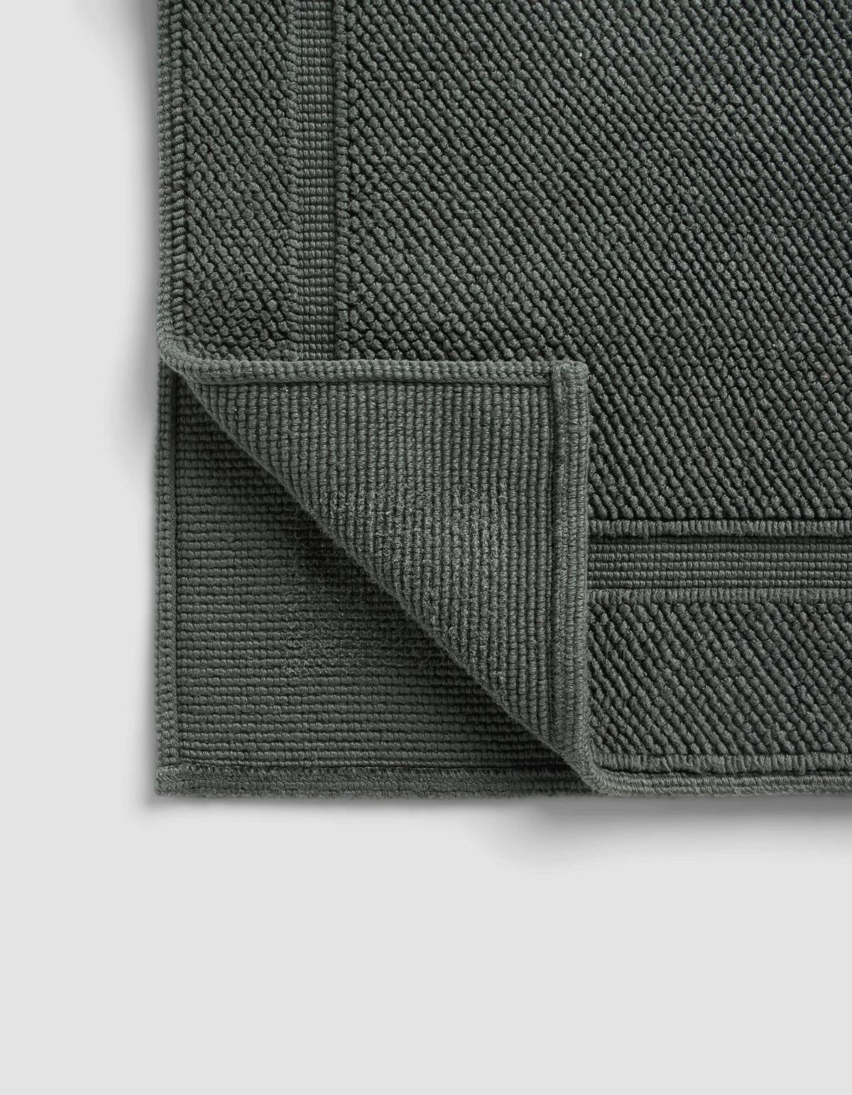 The Cozy Earth Classic Bath Mat, in dark gray with a textured woven pattern and bordered edges, is displayed on a light background with one corner folded to reveal the underside.
