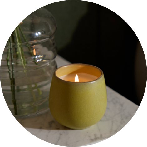 The Cozy Earth Bloom candle is lit and sitting on a marble surface next to a glass vase.