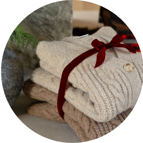 Two Cozy Earth Cable Knit Cardigans are folded and tied with a red ribbon near a vase with a plant.