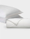 Two white pillows from Cozy Earth's Silk Insert Bundle are stacked diagonally behind a rolled-up white duvet with a visible tie closure, all set against a plain light gray background.|Filling:Standard