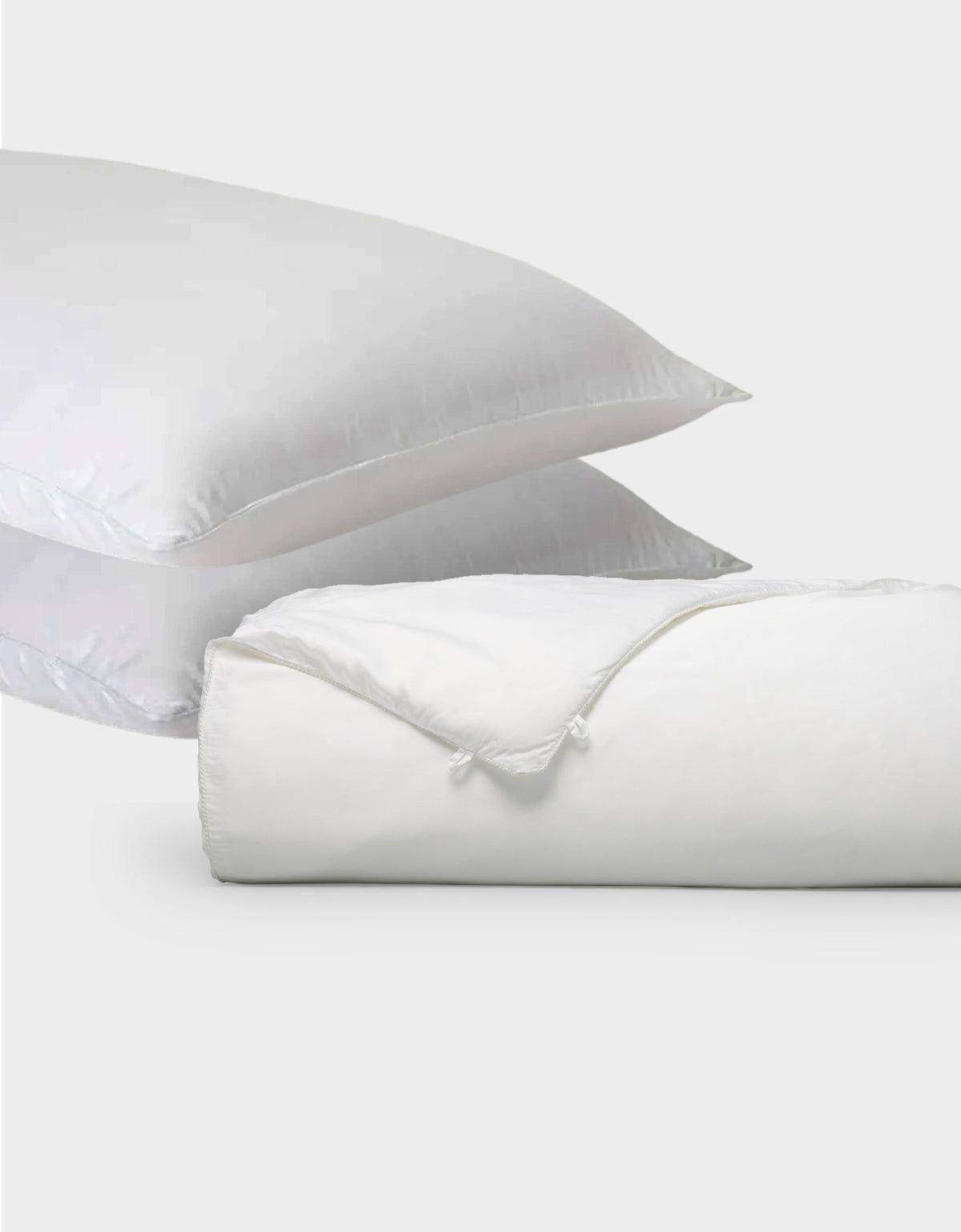 Two white pillows from Cozy Earth's Silk Insert Bundle are stacked diagonally behind a rolled-up white duvet with a visible tie closure, all set against a plain light gray background.|Filling:Standard