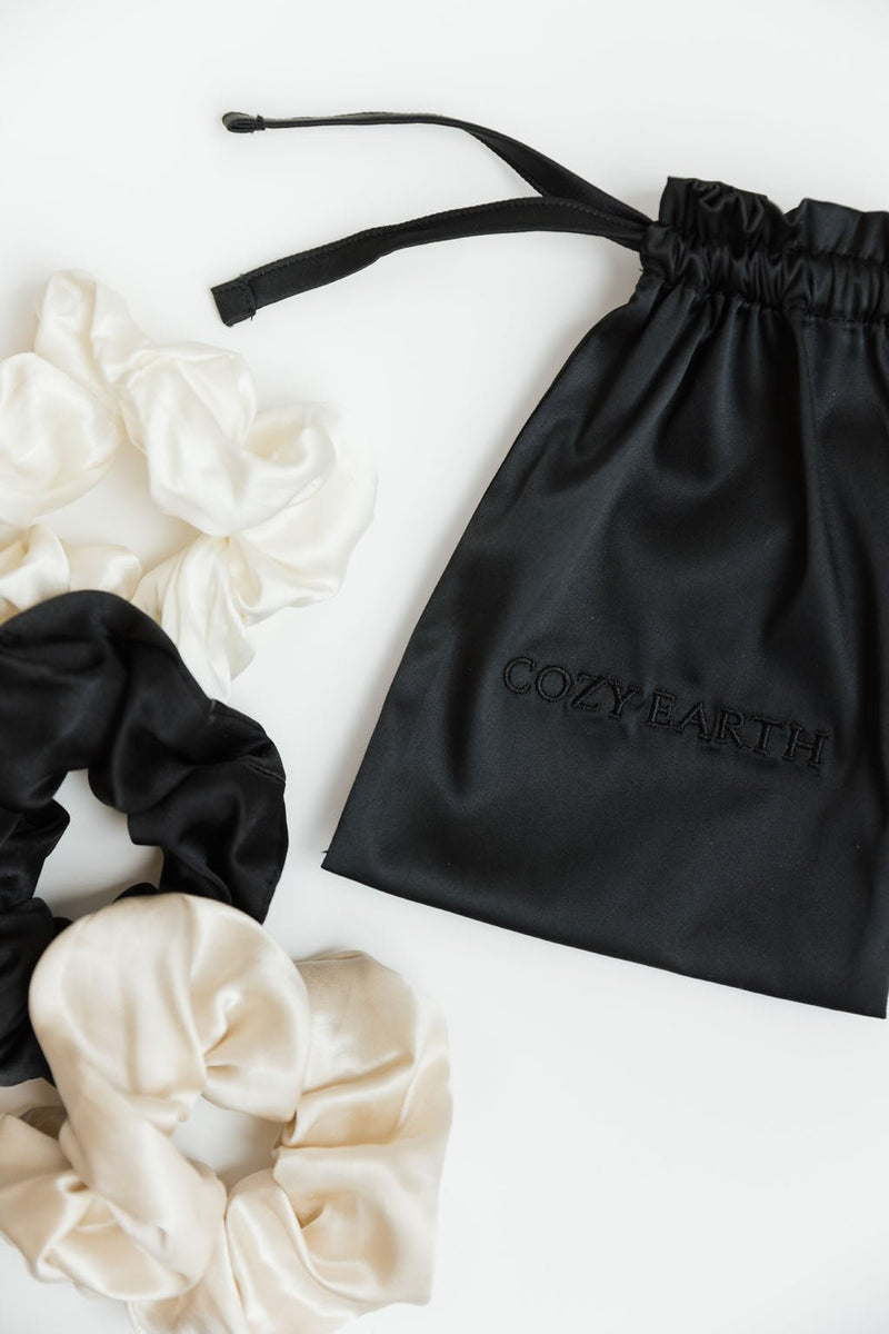A flat-lay image showcasing a black drawstring bag embroidered with "Cozy Earth" at the center. Surrounding the bag are three Silk Scrunchies from Cozy Earth: one black, one white, and one cream-colored, all crafted from a luxurious silky material. The background is a pristine white surface.