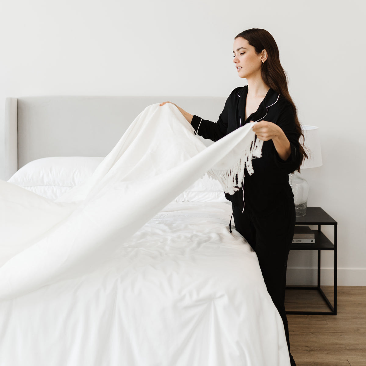 Woman in black pajamas laying white throw blanket over bed 