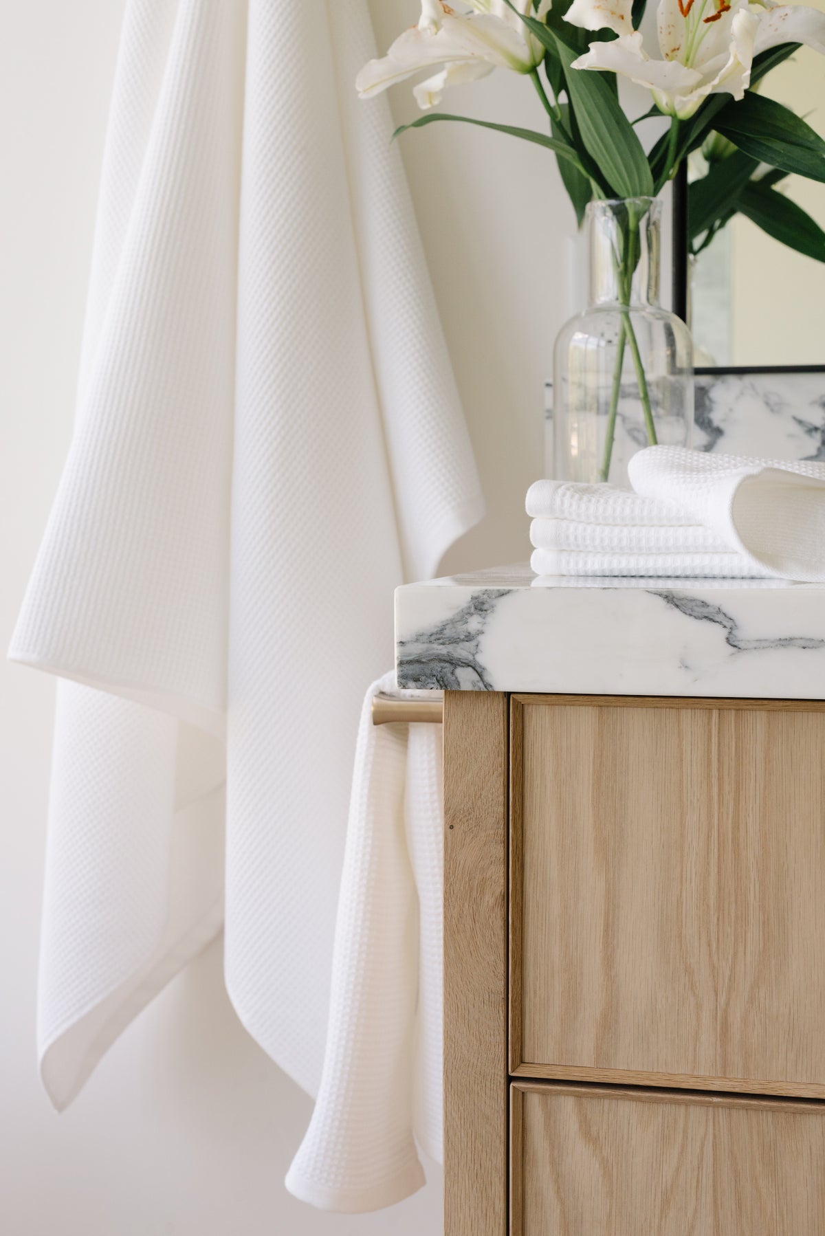 White Waffle Hand Towel hanging from a gold towel bar. The photo was taken at a white marble bathroom sink. A Waffle Bath Towel is seen hanging on the wall in the background. 