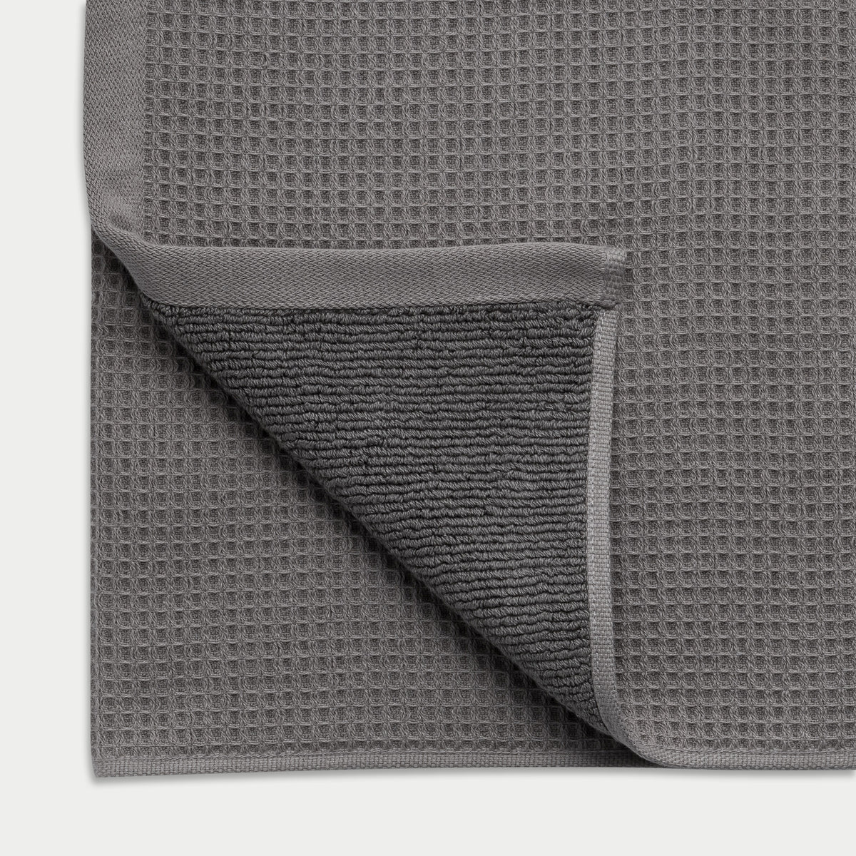 Charcoal Waffle Hand Towel resting flat on a white background. The corner is folded so that both sides of the towel can be seen. |Color:Charcoal